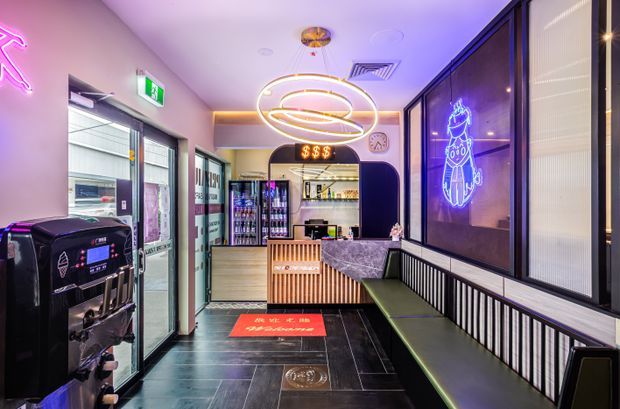 A compact, brightly lit commercial food-and-beverage venue interior with a takeaway counter, refrigerated drink displays, vending or service equipment, built-in banquette seating, neon wall graphics, dark tiled flooring, and a decorative ring pendant light. The space has a contemporary, playful, neon-accented fit-out with black, purple, pink, and warm timber tones.