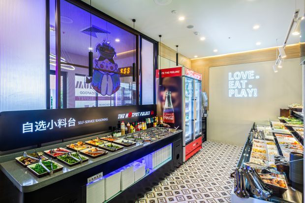 A brightly lit self-service food counter in a modern commercial eatery. Stainless-steel trays of prepared ingredients line a black counter beneath illuminated Chinese and English signage, with refrigerated drink displays, colourful graphic branding, patterned floor tiles, and a wall slogan reading “LOVE. EAT. PLAY!”.