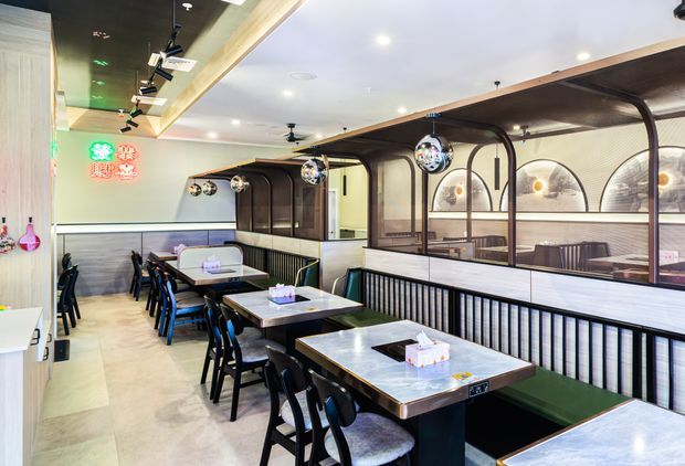A brightly lit contemporary restaurant interior with rows of booth seating and small square dining tables. Black metal-framed glazed partitions with arched details divide the booths, while hanging disco-ball pendant lights, track lighting, pale flooring, and dark upholstered seating create a playful modern dining atmosphere. Decorative illuminated signage and built-in banquettes are visible.