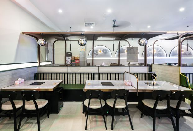A bright contemporary restaurant dining area with rows of marble-topped tables, black chairs, upholstered booth seating, and a decorative partition separating the dining space from a service or display area. The interior features curved dark framing, pendant globe lights, arched forms, pale wall finishes, and recessed ceiling lighting.
