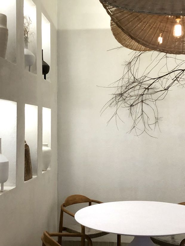 A softly lit dining nook with a round white table and curved wooden chairs. The space features white textured plaster walls, built-in recessed display niches, and a large woven natural-fibre pendant light with exposed branch-like decorative elements, creating an organic, serene atmosphere.