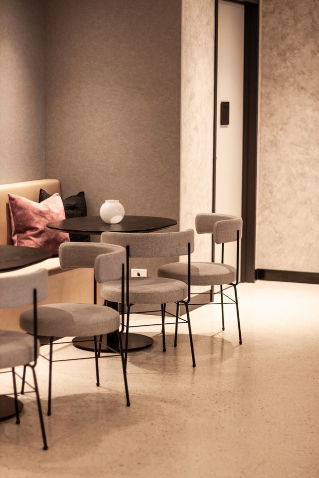 Warm, refined dining area with several compact round black dining tables and upholstered taupe chairs on a polished concrete floor. A built-in upholstered banquette with blush and dark cushions sits against textured taupe walls, while a full-height mirror or reflective panel and dark-framed doorway add depth. The space has a minimalist, contemporary hospitality-style fit-out with soft ambient lighting.