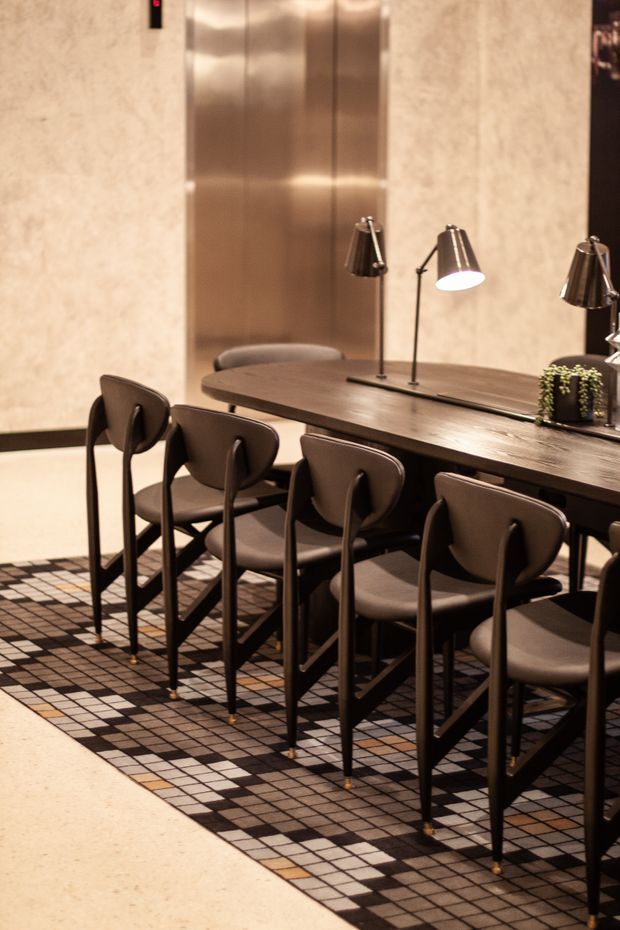 A refined dining area with a long dark timber dining table and multiple upholstered dining chairs. Two sculptural table lamps provide warm illumination, while a polished metallic elevator door and light stone-textured wall form the backdrop. The floor features a geometric black, grey, cream and beige tiled pattern, creating a sophisticated hospitality-like setting.
