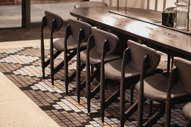 Close-up interior view of a dining area with a long dark timber dining table, multiple sculptural dark upholstered dining chairs, and a geometric black, white, and beige tiled floor. A glazed door or window and a partial brick wall are visible in the background, with warm ambient lighting and restrained contemporary detailing.