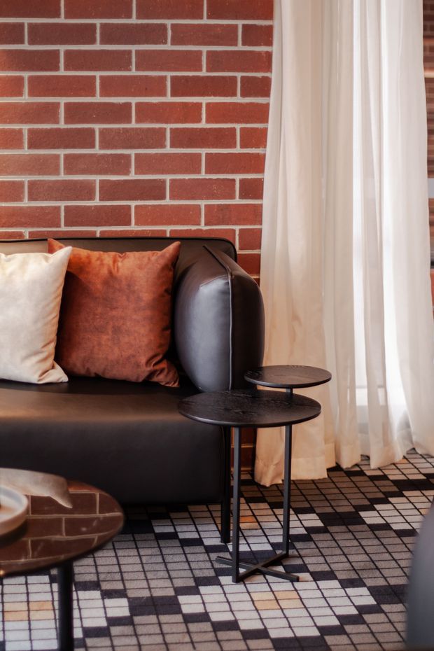 Close-up interior view of a stylish living room corner with a black leather sofa, rust and cream cushions, round black side tables, sheer white curtains, a red exposed-brick wall, and a bold black, white, and terracotta geometric rug. The space has a warm, graphic, contemporary feel.