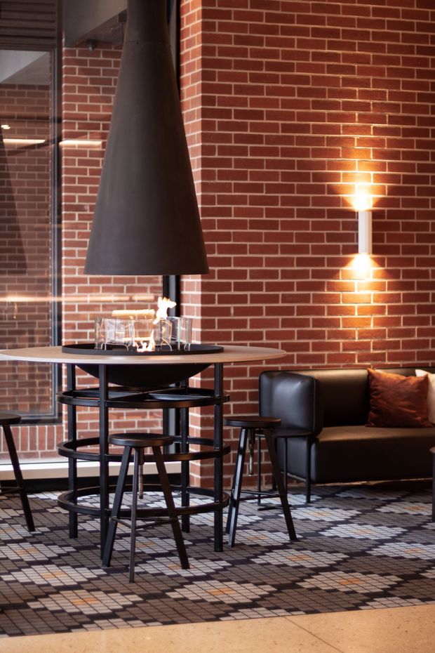 A warm, industrial-style lounge and dining area with exposed red brick walls, a suspended black cone fireplace over a round table, dark leather seating, black metal furniture, and a patterned tiled floor. A wall-mounted light provides a strong focal glow, while full-height glazing and brick reflections add depth.