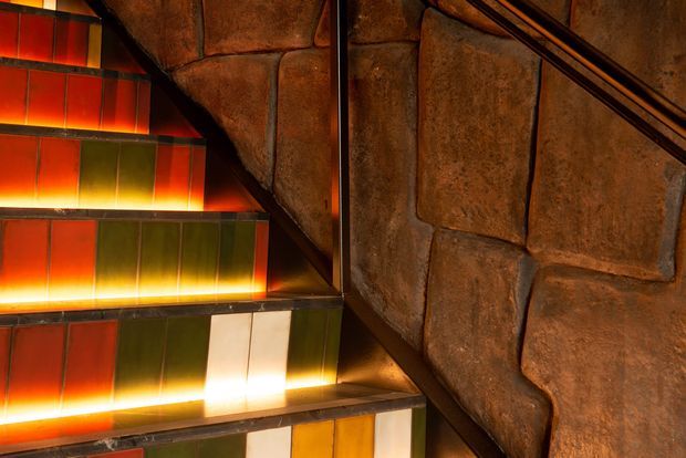 Close-up view of a dramatic stairway with translucent multicoloured panels illuminated from beneath, contrasted by a rough, sculptural brown stone or textured masonry wall and dark metal handrails. The warm orange, red, yellow and green lighting creates a moody, theatrical atmosphere.