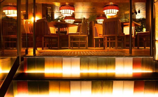 A warmly lit dining area viewed through large glazed panels, featuring substantial timber dining tables and high-backed timber chairs. Multiple decorative pendant lights create a dramatic amber glow, while reflective metal or glass surfaces and a raised threshold add visual depth. The image has the character of an intimate hospitality or restaurant interior.