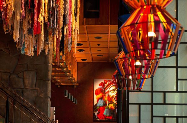 A dramatic, warm-toned interior featuring a sculptural staircase, dark stone-clad wall, richly timber-lined ceiling and cabinetry, colourful geometric pendant lights, textured hanging fibre art, and a vivid framed artwork. Large patterned glazing forms a strong architectural backdrop.