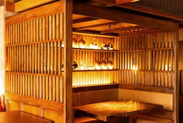 Warm, richly timber-lined dining or lounge booth with vertical slatted timber screens, exposed timber beams, integrated backlit shelving displaying decorative vessels, and a built-in upholstered banquette surrounding a substantial timber table. The space has intimate amber lighting and a strongly crafted, Japanese-inspired wood interior.