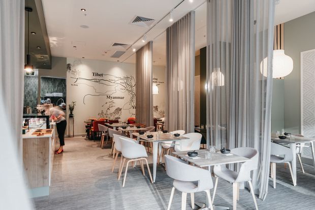 A bright contemporary restaurant or café interior with light timber-look flooring, pale walls, and a series of sheer floor-to-ceiling curtains dividing dining areas. White and light timber dining tables and chairs fill the space, with a service counter and kitchen area at left. A decorative wall graphic depicting a map and place names including Tibet and Myanmar forms a feature at the rear. Recessed and pendant lighting, exposed ceiling services, and reflective partitions create an airy, refined hospitality setting.