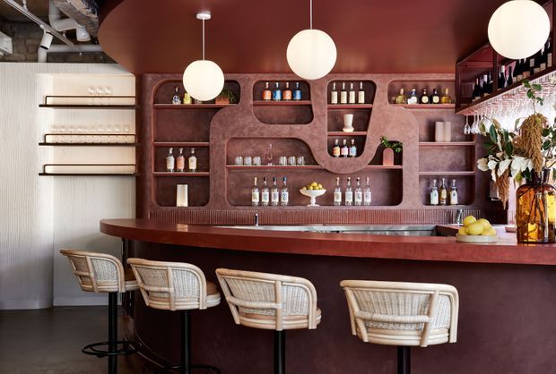 A stylish built-in home bar with a long dark burgundy counter and matching sculptural shelving, stocked with bottles and glassware. Four woven rattan bar stools line the counter, while globe pendant lights hang from a deep burgundy ceiling. Warm timber and rattan details contrast with textured plaster and dark flooring, creating a refined, moody hospitality-inspired interior.