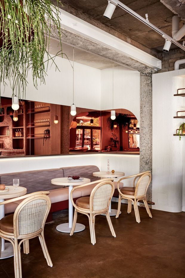 A warmly lit hospitality dining area with a built-in curved banquette, small round café tables, and woven rattan chairs. The space features a dark brown polished concrete floor, exposed rough concrete ceiling and column, white ribbed wall panelling, and an integrated timber-toned bar with open shelving and an arched mirror opening. Pendant lights, track lighting, and hanging greenery add visual interest. The overall atmosphere is refined, warm, and slightly retro.