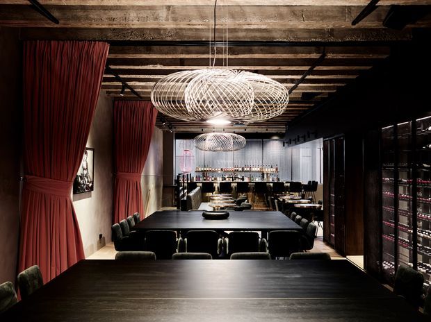 A dramatic, moody dining and entertaining space with long dark timber tables, deep upholstered seating, red floor-to-ceiling curtains, exposed timber ceiling beams, sculptural wire pendant lights, and a fully stocked bar at the rear. Dark built-in cabinetry and display shelving line the right wall, creating an intimate restaurant or home-bar atmosphere.