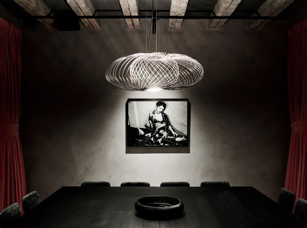 Dark, dramatic dining room with a long black dining table and matching chairs, framed black-and-white artwork, deep red curtains, a textured grey wall, and exposed concrete ceiling beams. A sculptural suspended loop pendant light forms the room's central feature, while a low round centrepiece sits on the table.