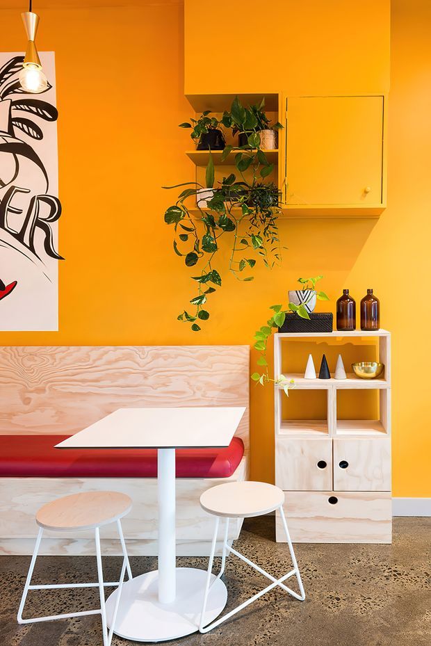 A vibrant, compact retro-inspired dining nook with saturated orange walls, a built-in pale timber bench with a red upholstered seat, and a white pedestal dining table surrounded by small stools and chairs. Open pale timber shelving and cabinetry display plants, bottles, and decorative objects, while a large black-and-white graphic artwork and pendant light add visual interest. The space has a playful, warm character and a terrazzo-like concrete floor.