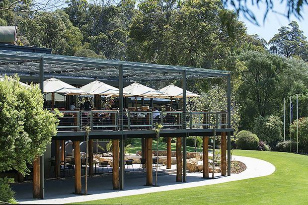 A two-level open-air pavilion or restaurant terrace set within a landscaped garden. The structure features a broad translucent or slatted roof, dark metal framing, timber posts, glass or metal balustrades, outdoor dining tables with umbrellas, and a covered lower seating area. Manicured lawn, curved paving, clipped shrubs, mature trees, and landscaped rockwork surround the building.