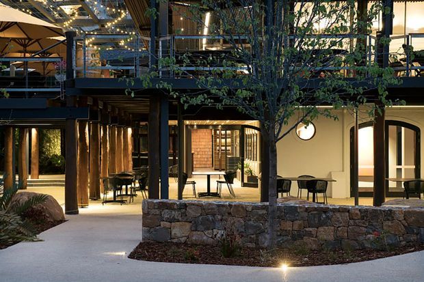 A landscaped courtyard or outdoor dining area within a multi-level commercial or hospitality building. The scene features dark steel posts and beams, timber columns, extensive glazing, warm interior lighting, outdoor tables and chairs, a mature tree, and a low rough-stone retaining wall. The architecture combines open-air circulation with sheltered terraces and planted areas, creating an inviting evening atmosphere.