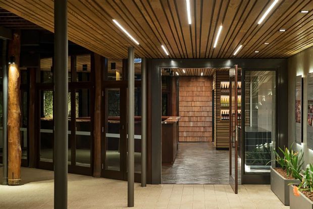 A warmly lit, refined interior passage or entry area with a slatted timber ceiling, dark metal structural posts, extensive dark-framed glazing and doors, and a tiled floor. The view leads toward a compact bar or display area with timber wall cladding and bottle shelving. Planters and natural timber details add an organic touch.