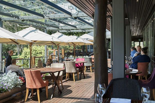 A bright, open-air restaurant terrace with timber decking, dining tables, upholstered chairs, large cream umbrellas, and a transparent glazed canopy supported by dark structural beams. The terrace connects to a glazed dining area on the right, with substantial timber posts and overhead timber slats creating a warm indoor-outdoor dining setting.
