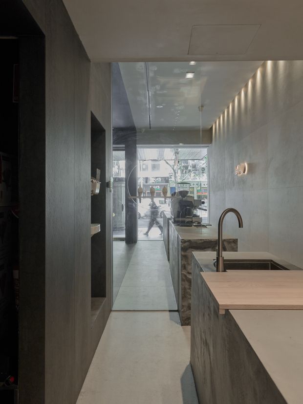 A narrow, sophisticated commercial interior viewed from behind a concrete-toned kitchen or service counter, looking toward a fully glazed street frontage. The space features a long pale concrete floor, textured grey wall surfaces, dark built-in shelving, a stone-faced island with an integrated stainless-steel sink and tall dark metal tap, and a pale timber benchtop extension. Recessed and linear wall lighting creates a moody, refined atmosphere, while pedestrians and neighbouring buildings are visible through the storefront glazing.