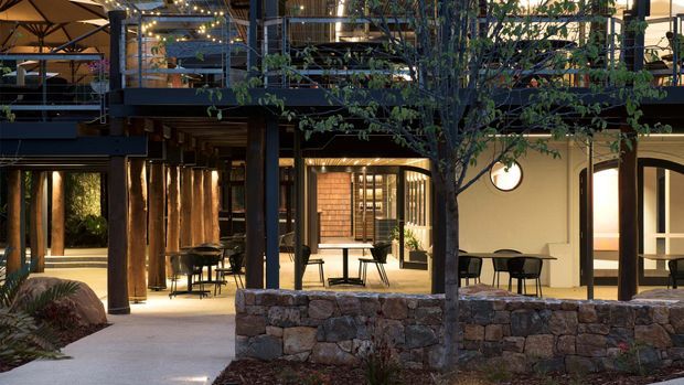 A warmly lit contemporary multi-level hospitality or commercial courtyard space viewed from outdoors. The building features dark steel structural framing, exposed timber soffits, upper-level terraces with planted greenery and string lights, arched openings, glazed doors, and a low rough-stone retaining wall in the foreground. Outdoor dining tables and chairs sit beneath a mature tree, creating an inviting evening atmosphere.