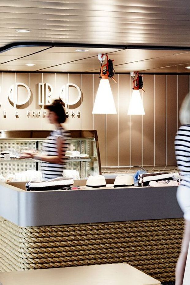 A stylish food-service counter inside a commercial café or restaurant. The space features a dark grey counter with thick woven rope-textured cladding, a refrigerated display case, warm beige vertically panelled walls, a ribbed ceiling, and suspended white pendant lights with exposed red-and-black fittings. Blurred staff or customers in striped clothing add a sense of activity. Large illuminated lettering on the rear wall identifies the venue as a food store or café.