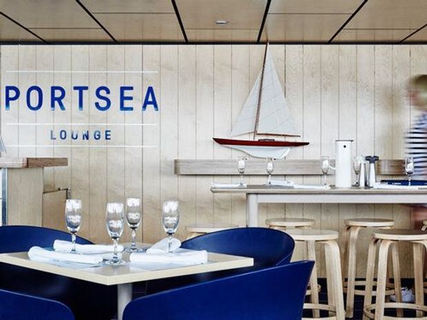 A stylish coastal-inspired lounge and dining area with deep blue upholstered chairs around a set dining table, pale timber wall panelling, and a compact bar or kitchenette with timber stools. A model sailboat and prominent blue “PORTSEA LOUNGE” signage reinforce the nautical theme.