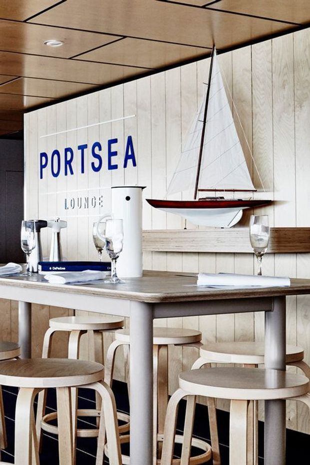 A compact coastal-themed hospitality lounge or café interior with a communal high table and multiple rounded wooden stools. The space features pale vertical timber wall panelling, a warm timber-panel ceiling with dark linear seams, a dark tiled lower wall, and a long wooden shelf. Blue 'PORTSEA LOUNGE' lettering and a mounted model sailboat reinforce the nautical character. Glassware and table settings are arranged on the table.