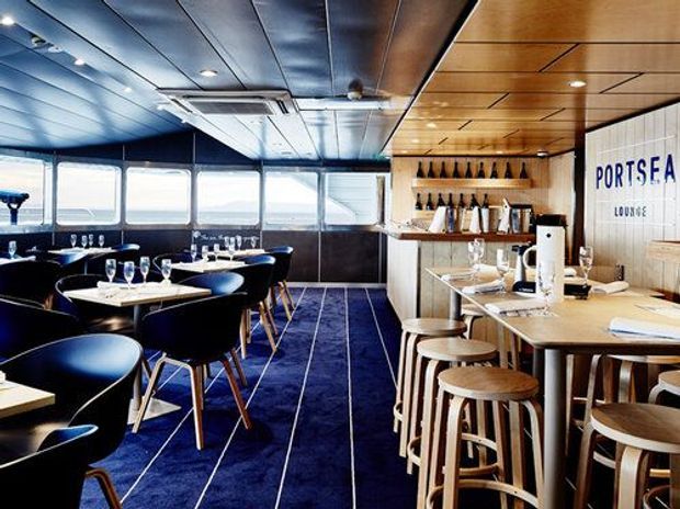 Interior view of a waterfront-style restaurant or lounge with extensive glazing, dark blue carpet, navy upholstered dining chairs, timber ceiling panels, and a timber service bar with shelving and stools. A prominent wall sign reads “PORTSEA LOUNGE.”