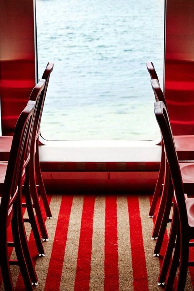 A symmetrical, stylised interior view of a compact passenger cabin or transport lounge, with rows of glossy dark red seating framing a large rounded rectangular window. The floor has bold alternating red and beige stripes, while red wall panels and a pale window surround create a strong graphic, high-contrast composition.