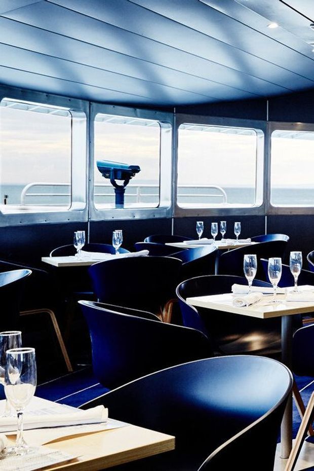 A refined dining room aboard a vessel, with dark navy upholstered chairs, neatly set white tables, and large metal-framed windows overlooking the ocean. The space has a streamlined marine-inspired interior with a paneled ceiling, dark wall lining, and cool blue-toned lighting.