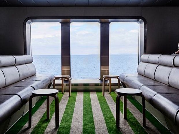A compact, highly stylised lounge interior with opposing light-grey upholstered bench sofas, small round black tables, and three large rounded-rectangle windows overlooking open water. The room has dark walls and ceiling, metallic-toned framing, and a bold striped green-and-grey carpet, creating a futuristic marine or transport-like atmosphere.