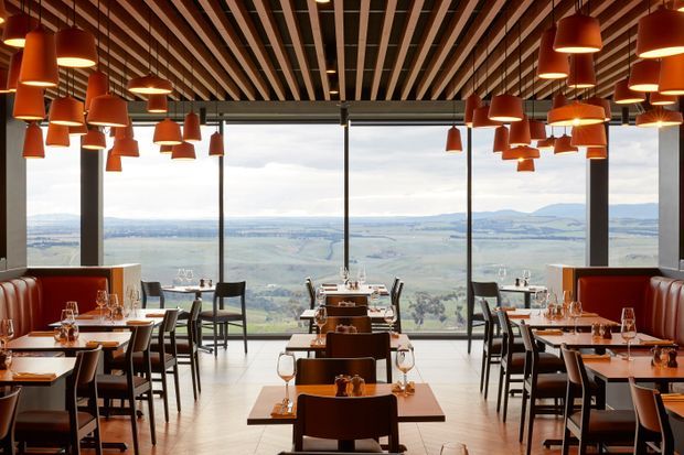 A sophisticated restaurant dining room with rows of tables, upholstered banquette seating, dark timber dining chairs, and extensive floor-to-ceiling glazing overlooking a broad rural landscape. The ceiling features a striking slatted timber grid with numerous suspended terracotta pendant lights, creating a warm, dramatic hospitality interior.