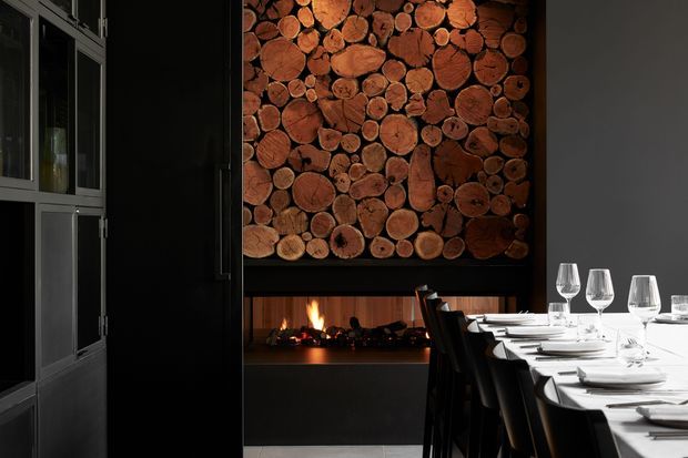 A dramatic contemporary dining room with a long formally set dining table, dark cabinetry or glazed joinery, and a linear fireplace beneath a large feature wall made from stacked cut timber logs. The room uses strong black and charcoal tones contrasted with warm natural wood and firelight, creating a refined, moody atmosphere.