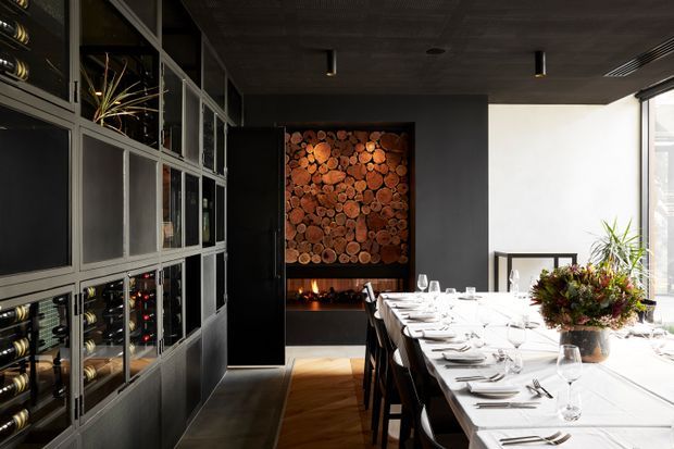 A sophisticated dining room with a long formally set dining table, dark built-in wine storage and display cabinetry along the left wall, and a dramatic feature wall of stacked timber end-grain logs above a linear fireplace. The room has dark charcoal walls and ceiling, warm timber flooring, black-framed glazing, and strong natural light from the right. The styling is refined and contemporary with a warm, intimate atmosphere.