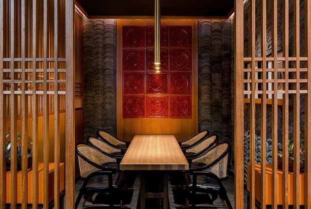 A richly detailed dining room viewed through timber screens, featuring a long solid-wood dining table surrounded by woven-back dark chairs. Warm timber wall and ceiling finishes, patterned dark textured wall panels, a red decorative artwork grid, and a central pendant light create an intimate, dramatic atmosphere. Vertical timber slats and latticework frame the room on both sides.