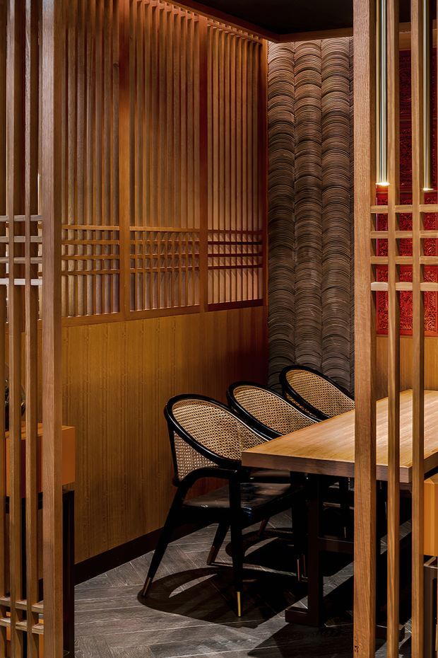 A richly detailed dining area viewed through vertical timber screens. The space features warm timber wall panelling and latticework, a dark patterned feature wall with repeating curved relief, a timber dining table, and three black-framed woven cane chairs. Dark herringbone flooring and red decorative wall panels add contrast, creating an intimate, sophisticated interior with strong Japanese-inspired detailing.