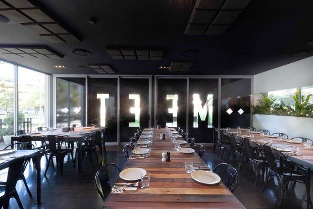 A contemporary restaurant dining room with multiple long timber tables set with white plates, glassware, and cutlery. Black chairs and dark flooring contrast with a black ceiling featuring geometric recessed panels and exposed circular fixtures. Full-height glazed doors and windows bring in daylight, while a dark glazed rear partition carries large illuminated lettering and a raised planter with greenery runs along the right side.