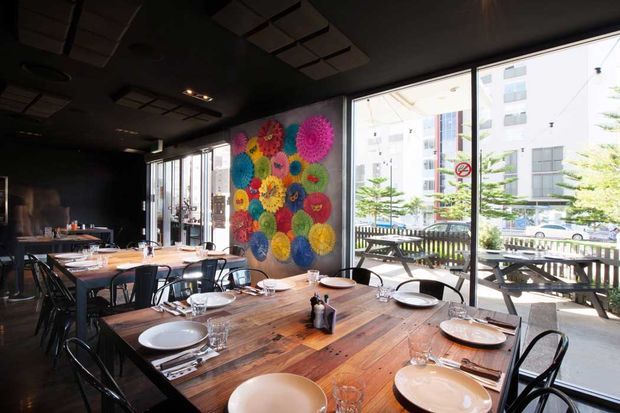 A dark, contemporary restaurant dining interior with long rustic timber tables, black metal chairs, table settings, and large floor-to-ceiling windows opening to an outdoor terrace. A colourful circular artwork forms a vibrant feature wall, while acoustic ceiling panels and black finishes create a dramatic, urban atmosphere.