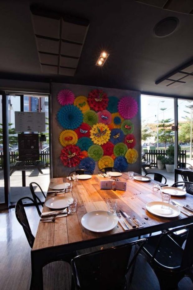 A dark, contemporary dining room in a restaurant or hospitality setting, centered on a large rustic timber communal dining table set with white plates, cutlery, and glassware. Black chairs surround the table, while floor-to-ceiling windows provide abundant daylight. A colorful wall installation of paper-like floral rosettes and theatrical masks forms a vivid feature backdrop. The ceiling is dark with recessed lighting and a circular air vent.