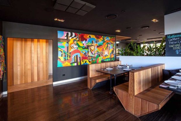 A dark, contemporary restaurant or hospitality dining space with rich timber flooring, timber-clad partitions and banquette seating, a dark ceiling with recessed lighting, and a large colourful mural as the main feature wall. Dining tables, place settings, and a chalkboard menu are visible, with greenery behind a partial divider.