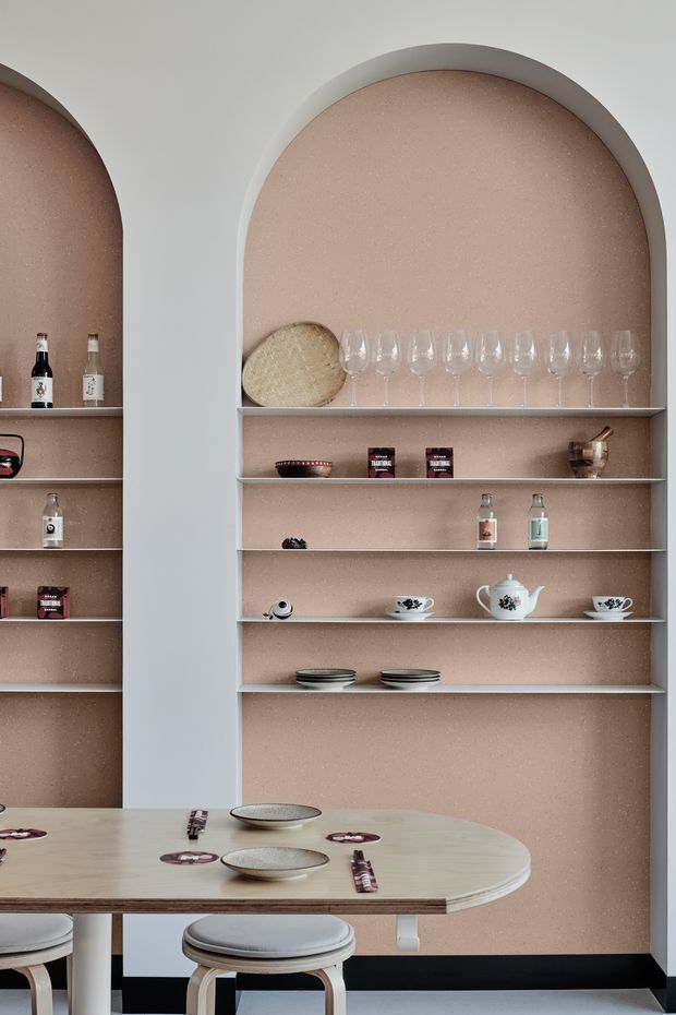 A refined dining area with a rounded rectangular dining table and stools positioned before built-in arched display shelving. The shelving has a dusty blush-pink textured backing, pale grey-white framing and shelves, and displays glassware, crockery, bottles, and decorative objects. The room has a soft neutral palette with dark skirting and a contemporary, gently retro character.