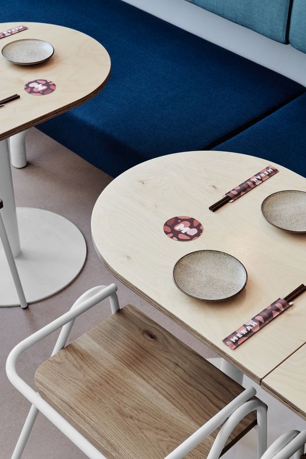 Close-up view of a contemporary dining or café-style seating area. Pale plywood tables with rounded corners are set with ceramic bowls and patterned placemats, paired with a dark navy upholstered bench and a light wood-and-white tubular-frame chair. The space has a restrained, graphic palette with soft pink flooring and clean modern detailing.