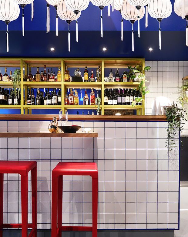 A lively, compact bar interior with a deep navy-blue ceiling and upper wall, white globe-style pendant lanterns, and illuminated lime-yellow open shelving displaying liquor bottles. The bar front and backsplash are clad in white square tiles with dark grout, topped by timber counters. Bright red stools provide a bold accent, while trailing greenery softens the highly graphic, colourful scheme.