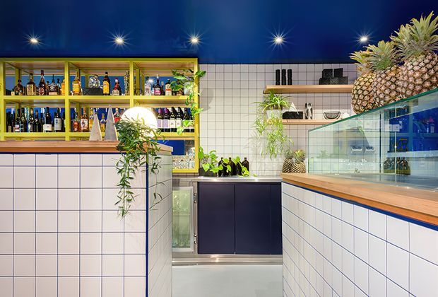 A vibrant contemporary commercial bar or hospitality interior with deep blue ceilings, white square tiled walls and counters, yellow-framed open bottle shelving, dark blue cabinetry, timber-edged glass display cases, and abundant plants. Bottles and pineapples are displayed throughout, with pendant or starburst ceiling lights adding a dramatic nightlife atmosphere.