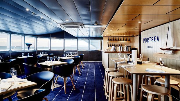 A stylish waterfront dining and bar interior with panoramic windows, dark blue upholstered seating, a deep blue carpet, a timber-lined ceiling, and a light timber bar counter with stools. Built-in bottle shelving and nautical detailing, including a small suspended boat, create a refined contemporary maritime atmosphere.