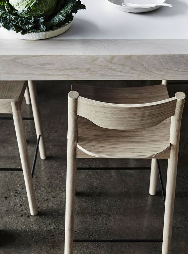 Close-up product-style image of a light timber dining table and matching wooden chairs, showing curved slatted chair backs, pale wood legs, and a dark textured floor. A white countertop or tabletop with a ceramic bowl and leafy greens is visible at the top of the frame.