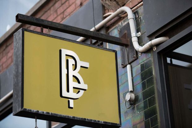 Close-up exterior architectural detail of a projecting business sign mounted beside a doorway. The sign has a mustard-yellow face with a bold black-and-white monogram, enclosed in a dark metal frame. Behind it is an urban facade combining exposed red brick, dark painted panels, glossy dark green rectangular tiles, visible white service pipes, and a black-framed glazed entrance.