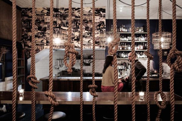 A dimly lit, atmospheric bar or restaurant interior viewed through a dramatic screen of thick knotted rope. The space features a dark timber counter and tables, bar shelving stocked with bottles, white subway-tile wall sections, black-and-white photographic wall imagery, pendant or candle-like accent lighting, and patrons seated at the bar. The overall look is eclectic and industrial with a strong nautical or theatrical character.
